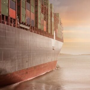 A colossal cargo ship loaded with containers navigates through calm waters against a vibrant sunset sky.